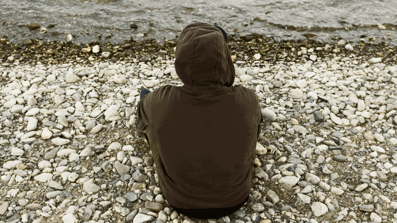 Ein junger Mann sitzt mit dem Rücken zur Kamera an einem Flußufer. Er trägt eine Shirt mit einer Kapuze. Ein junger Mann sitzt mit dem Rücken zur Kamera an einem Flußufer. Er trägt eine Shirt mit einer Kapuze.