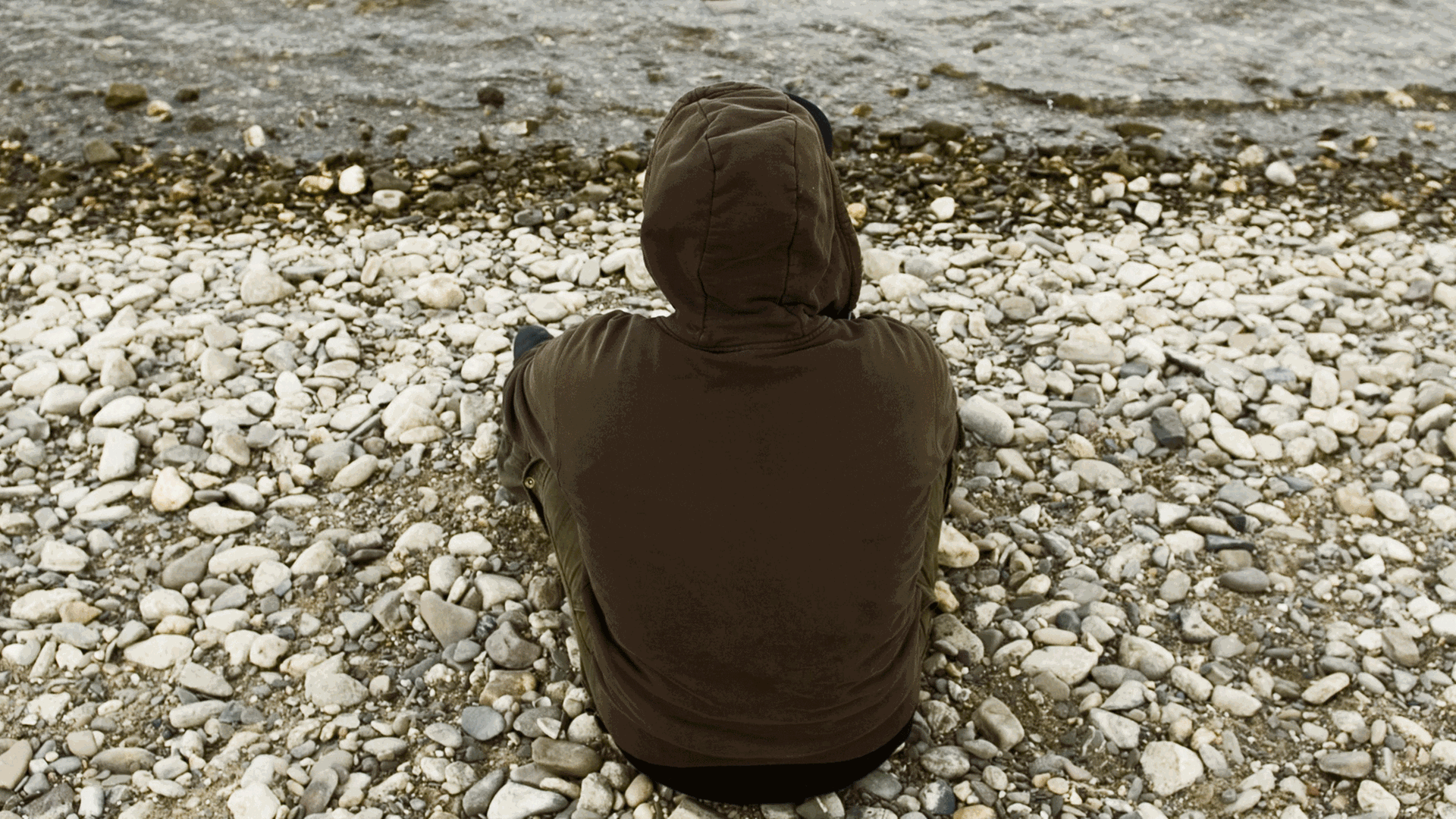 Ein junger Mann sitzt mit dem Rücken zur Kamera an einem Flußufer. Er trägt eine Shirt mit einer Kapuze.