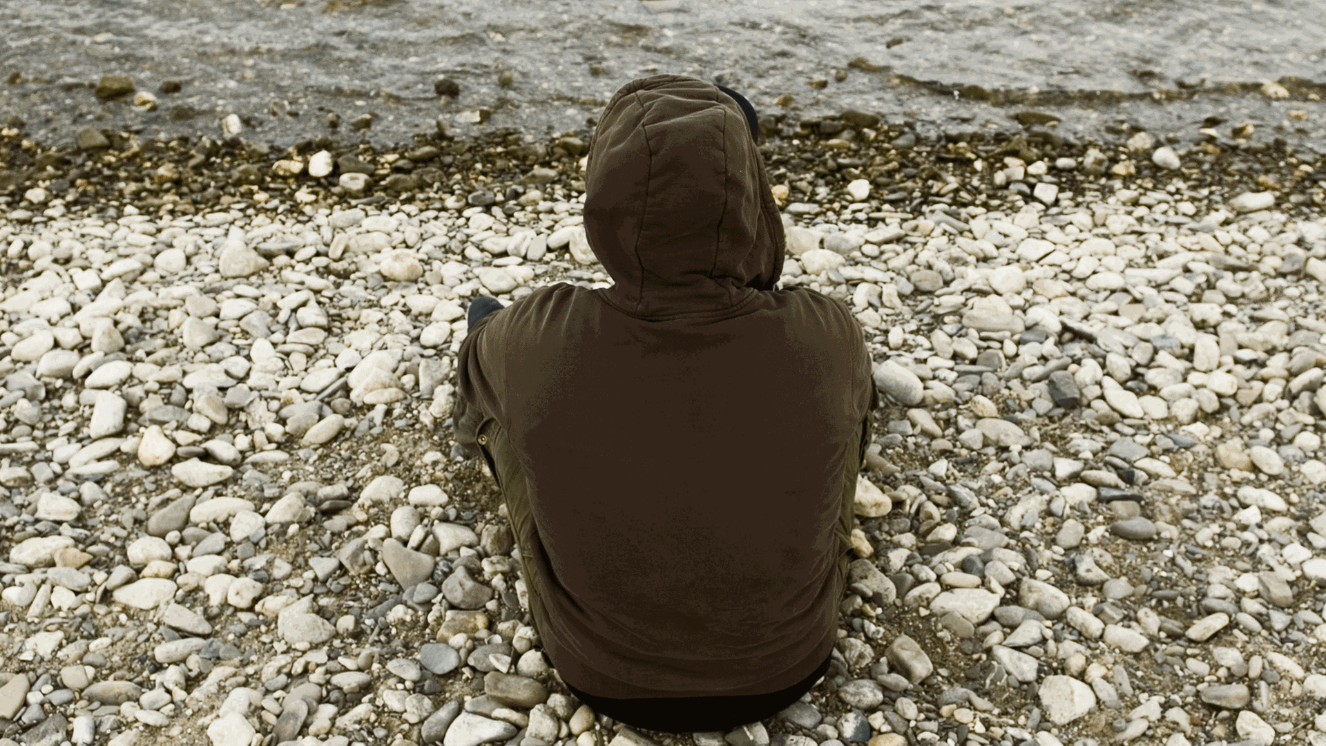 Ein junger Mann sitzt mit dem Rücken zur Kamera an einem Flußufer. Er trägt eine Shirt mit einer Kapuze. Ein junger Mann sitzt mit dem Rücken zur Kamera an einem Flußufer. Er trägt eine Shirt mit einer Kapuze.
