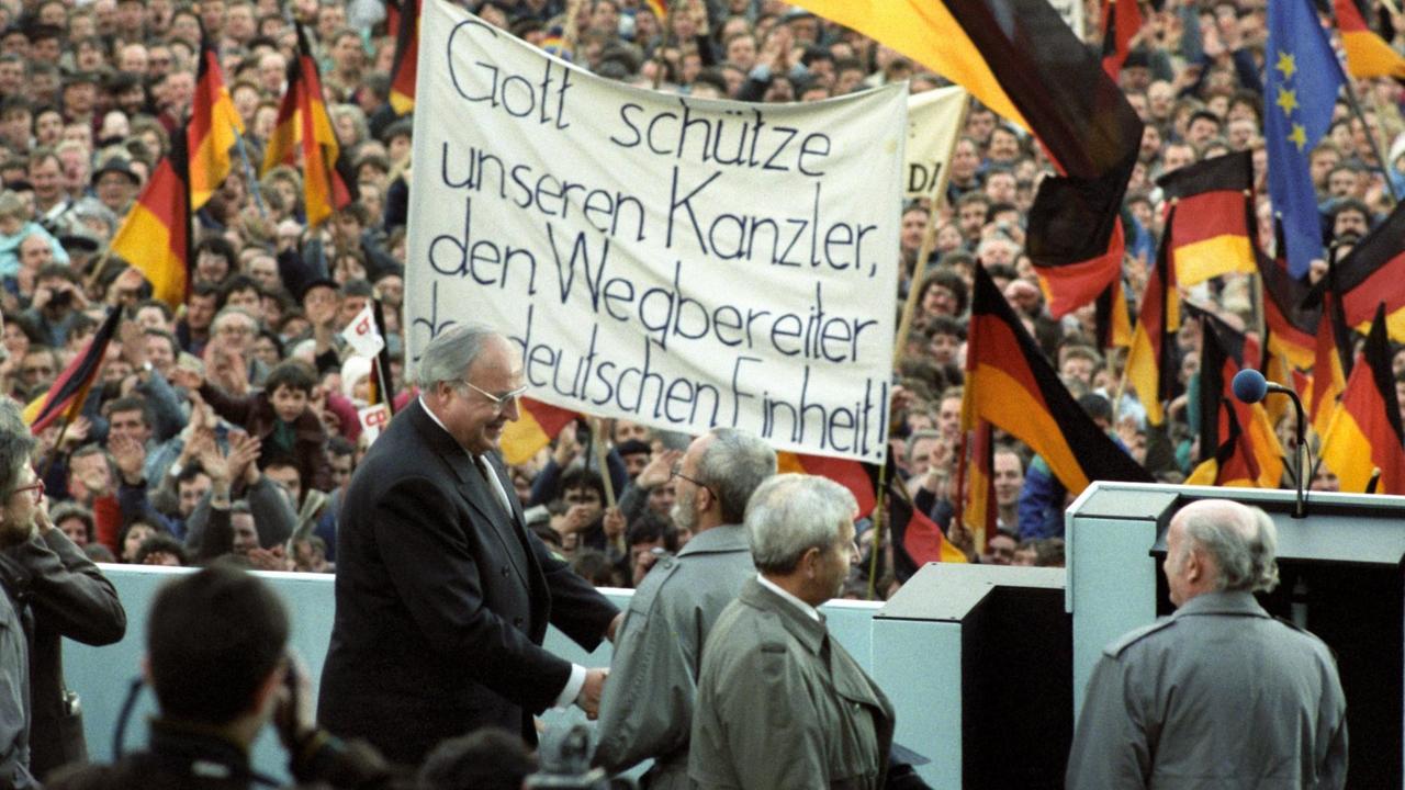 Bundeskanzler Helmut Kohl vor einer Menschenmenge mit Deutschland-Flaggen und deinem Spruchband: "Gott schütze unseren Kanzler, den Wegbereiter der deutschen Einheit"