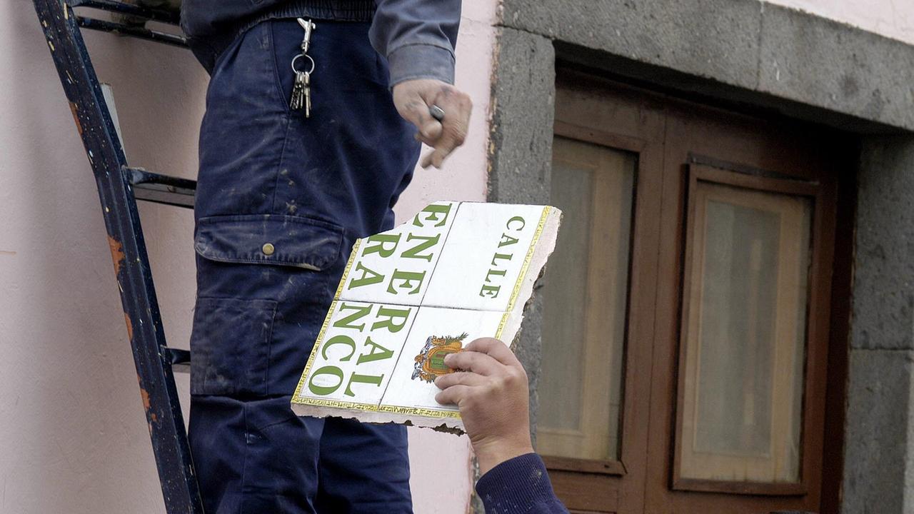 Arbeiter wechseln ein Straßenschild aus, auf dem "Calle General Franco" steht, ein Arbeiter steht auf einer Leiter, nur seine Beine sind sichtbar, ein anderer nimmt das Schild in die Hand Arbeiter wechseln ein Straßenschild aus, auf dem "Calle General Franco" steht, ein Arbeiter steht auf einer Leiter, nur seine Beine sind sichtbar, ein anderer nimmt das Schild in die Hand