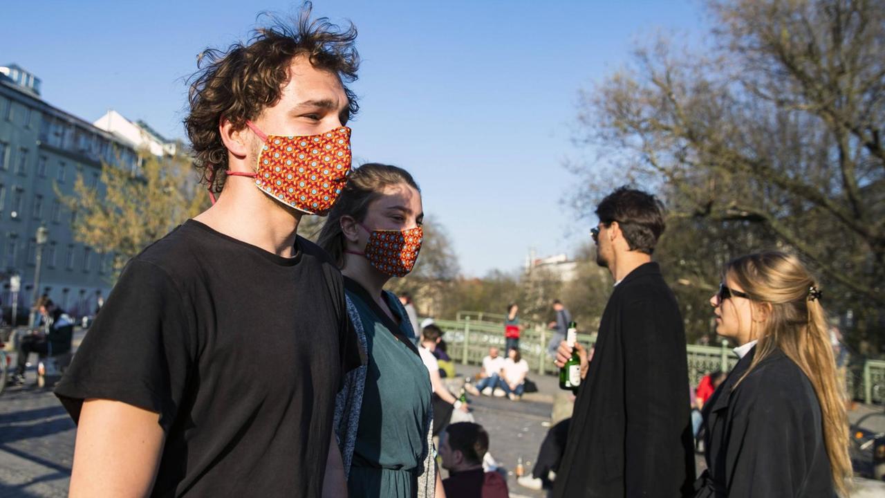 Ein junger Mann und eine junge Frau tragen selbstgemachte bunte Mundschutzmasken. Ihnen kommen ein Mann und eine Frau ohne Mundschutzmaske entgegen. Ein junger Mann und eine junge Frau tragen selbstgemachte bunte Mundschutzmasken. Ihnen kommen ein Mann und eine Frau ohne Mundschutzmaske entgegen.