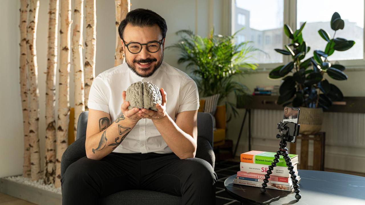 Umut Özdemir sitzt auf einer Couch, in der Hand eines Modells von einem Gehirn. Umut Özdemir sitzt auf einer Couch, in der Hand eines Modells von einem Gehirn.