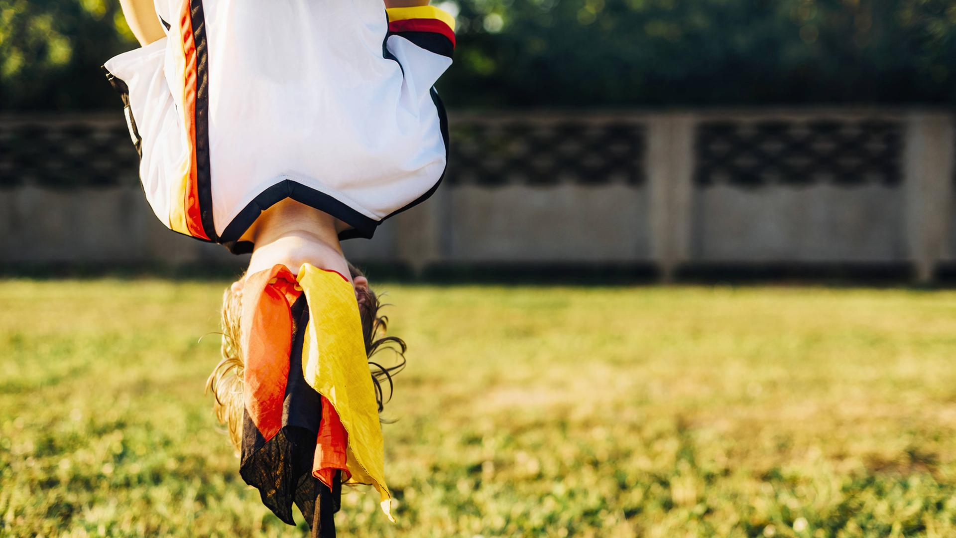 Ein Junge, der ein Trikot der deutschen Fußball-Nationalmannschaft trägt, hängt mit dem Kopf nach unten über einer Wiese. Über seinem Gesicht hängt die deutsche Flagge in Falten. Ein Junge, der ein Trikot der deutschen Fußball-Nationalmannschaft trägt, hängt mit dem Kopf nach unten über einer Wiese. Über seinem Gesicht hängt die deutsche Flagge in Falten.