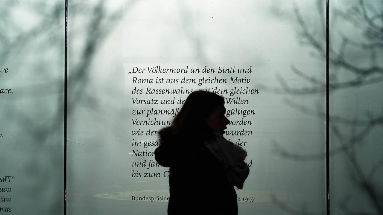 Die Silhouette einer Frau zeichnet sich ab vor einer Tafel mit Texten zum Völkermord an den Sinti und Roma. Die Silhouette einer Frau zeichnet sich ab vor einer Tafel mit Texten zum Völkermord an den Sinti und Roma.