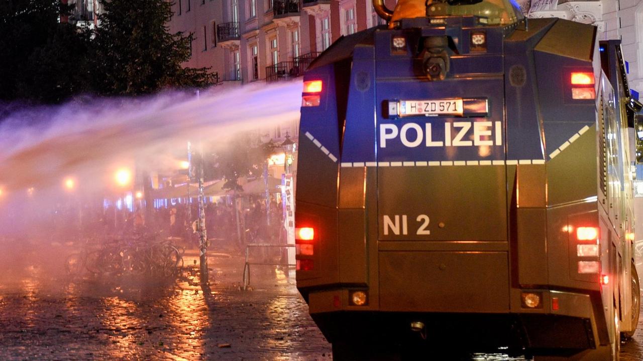 Das Bild zeigt einen Wasserwerfer in der Nacht von hinten, der gerade seitlich Wasser gegen einen Demonstranten verspritzt. Das Bild zeigt einen Wasserwerfer in der Nacht von hinten, der gerade seitlich Wasser gegen einen Demonstranten verspritzt.