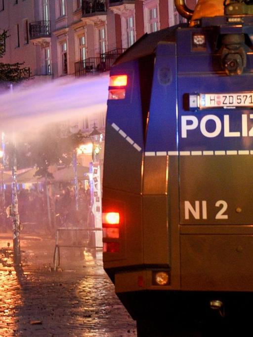Das Bild zeigt einen Wasserwerfer in der Nacht von hinten, der gerade seitlich Wasser gegen einen Demonstranten verspritzt.