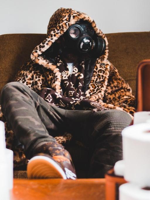Ein junger Mensch sitzt maskiert mit einer Gasmaske auf einer Couch und blickt ins off, während sich auf dem Tisch das Klopapier stapelt.