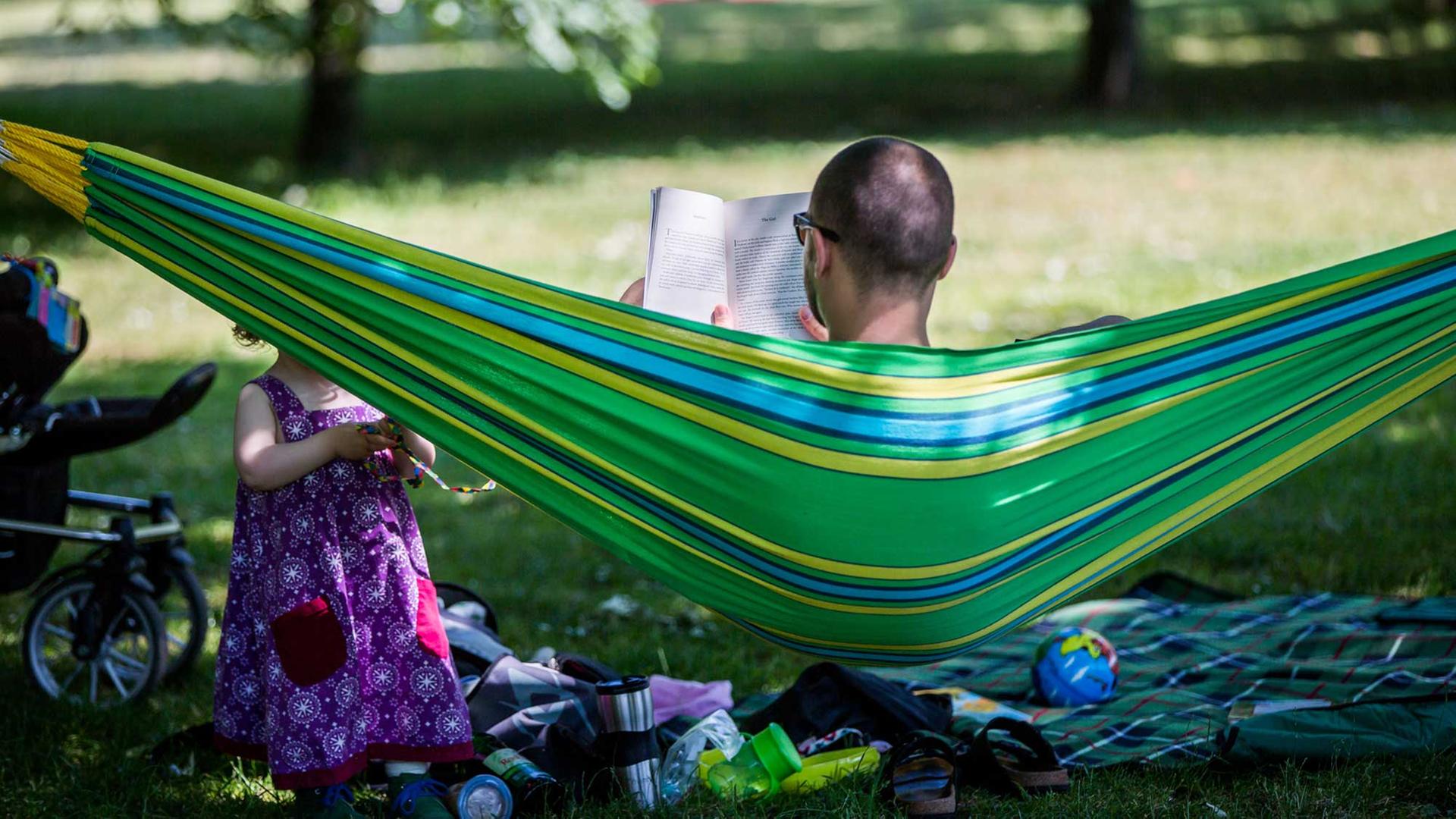 Ein Vater liest in Frankfurt am Main ein Buch in einer Hängematte im Günthersburgpark. Vor ihm spielt seine kleine Tochter im Schatten der Bäume. Ein Vater liest in Frankfurt am Main ein Buch in einer Hängematte im Günthersburgpark. Vor ihm spielt seine kleine Tochter im Schatten der Bäume.