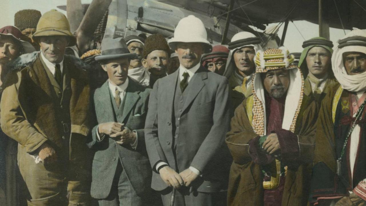 Ein historisches Farbfoto zeigt mehrere britische Kolonialbeamte neben Männern in arabischer Tracht vor einem Propellerflugzeug stehend