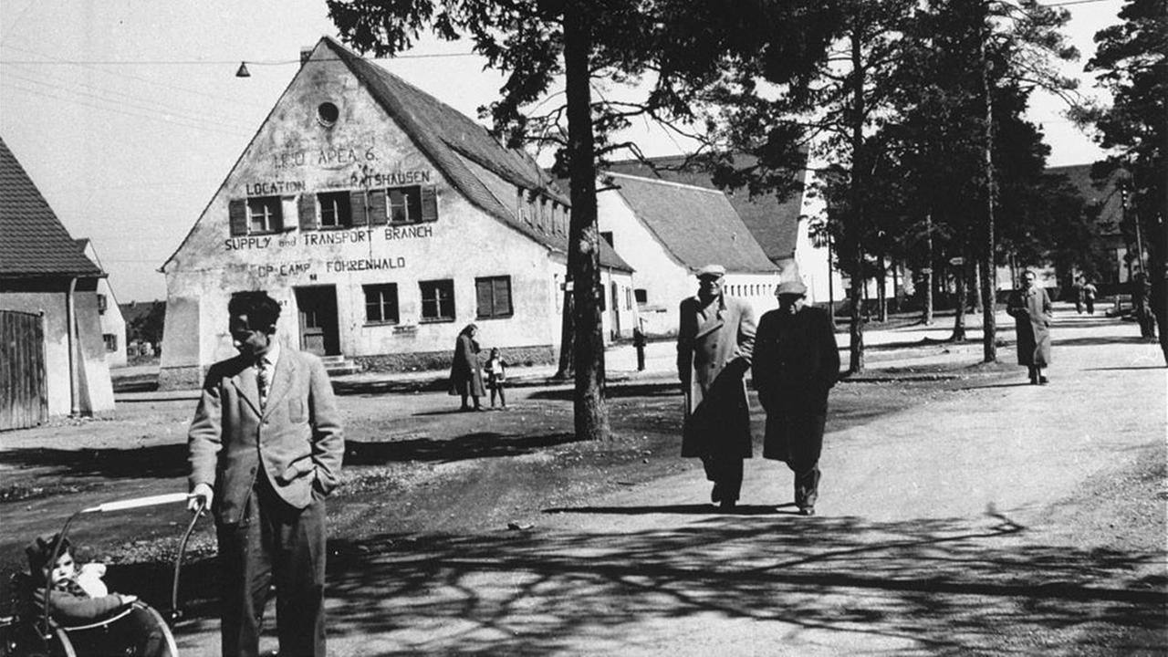 Das Schwarz-Weiß-Bild zeigt eine Straßenszene im DP-Lager Föhrenwald in Bayern mit mehreren Menschen, im Hintergrund Bäume und Gebäude. Das Schwarz-Weiß-Bild zeigt eine Straßenszene im DP-Lager Föhrenwald in Bayern mit mehreren Menschen, im Hintergrund Bäume und Gebäude.