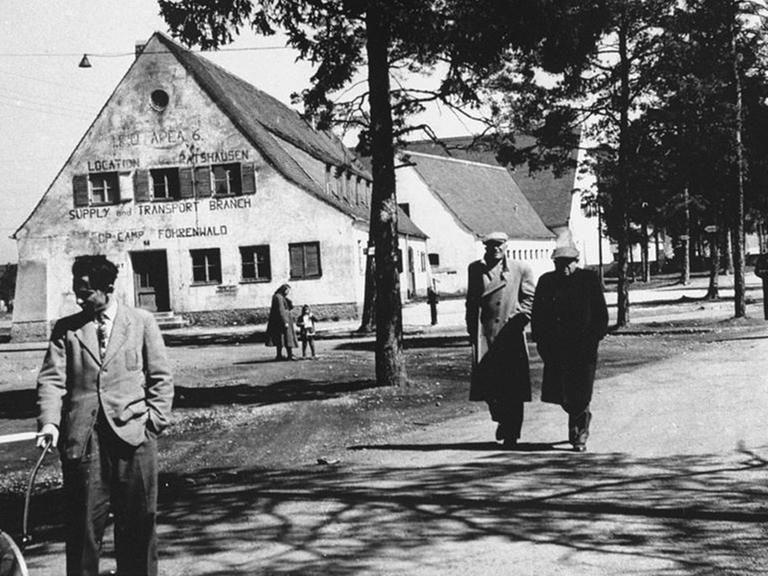 Das Schwarz-Weiß-Bild zeigt eine Straßenszene im DP-Lager Föhrenwald in Bayern mit mehreren Menschen, im Hintergrund Bäume und Gebäude.