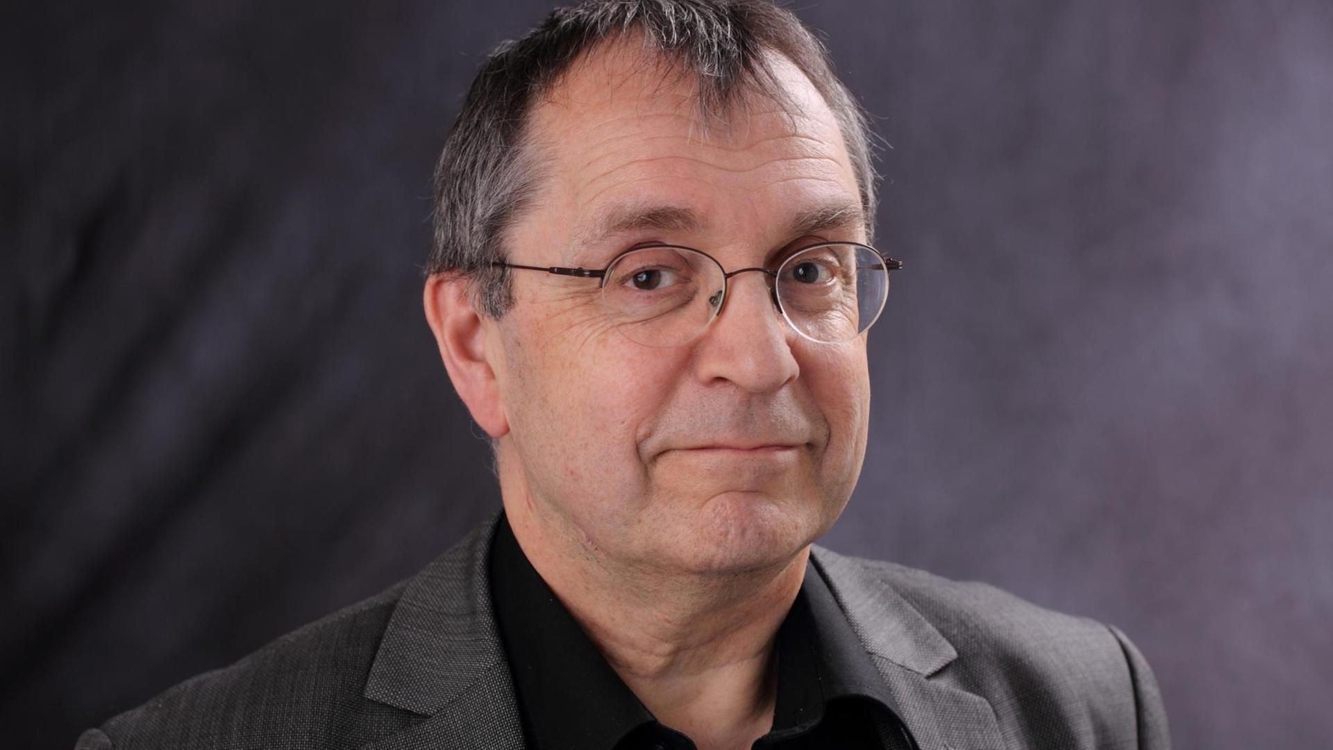 Ein Mann mit graumeliertem Haar und Brille steht vor einem grauen Hintergrund. Ein Mann mit graumeliertem Haar und Brille steht vor einem grauen Hintergrund.