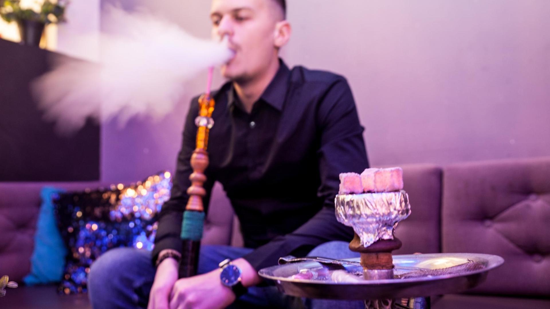 Ein junger Mann sitzt in einer lila illuminierten Shisha-Bar und atmet Dampf aus. Das Bild ist leicht unscharf und vermittelt dadurch eine leicht schummrige Atmosphäre. Ein junger Mann sitzt in einer lila illuminierten Shisha-Bar und atmet Dampf aus. Das Bild ist leicht unscharf und vermittelt dadurch eine leicht schummrige Atmosphäre.