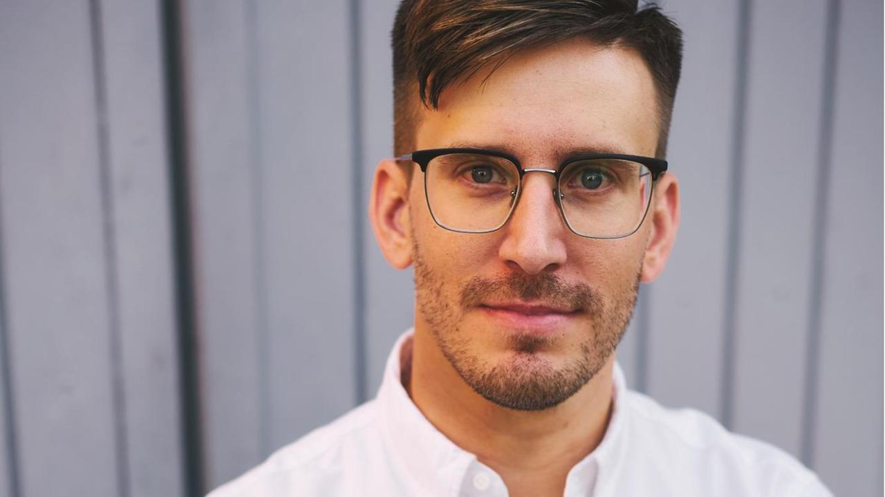 Sebastian Ostritsch, mit Brille, im offenen weißen Hemd, steht vor einer grauen Holzwand. Sebastian Ostritsch, mit Brille, im offenen weißen Hemd, steht vor einer grauen Holzwand.