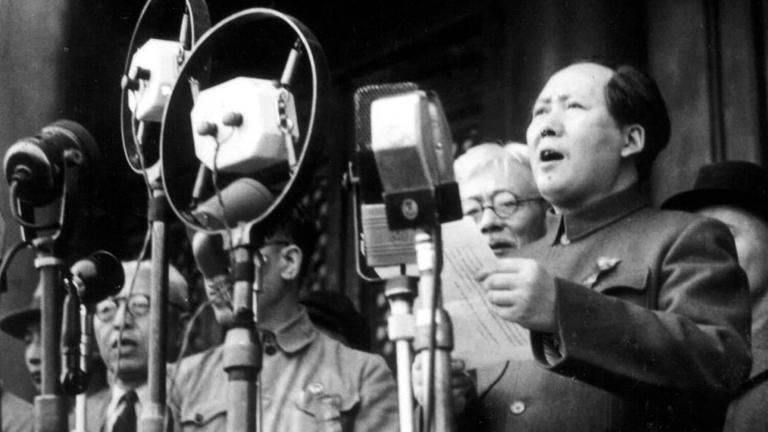 Mao Zedong steht in Uniform vor einem Mikrofon und verkündet mit einem Papier in der Hand die Chinesische Volksrepublik. Um ihn herum stehen Funktionäre. 