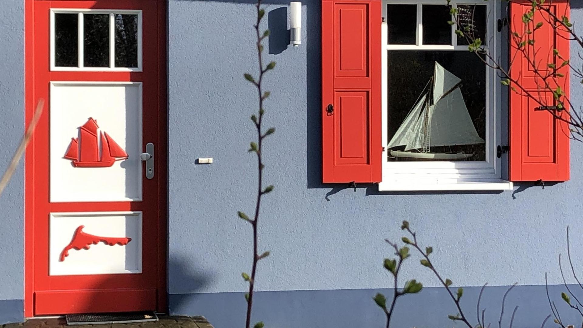 Auf dieser modernen Variante einer Darßer Tür ist die Silhouette eines Segelschiffs und ein Inselumriss zu sehen. Das Haus ist grau gestrichen, links und rechts vom Fenster sind rote Holzfensterläden. Auf dieser modernen Variante einer Darßer Tür ist die Silhouette eines Segelschiffs und ein Inselumriss zu sehen. Das Haus ist grau gestrichen, links und rechts vom Fenster sind rote Holzfensterläden.