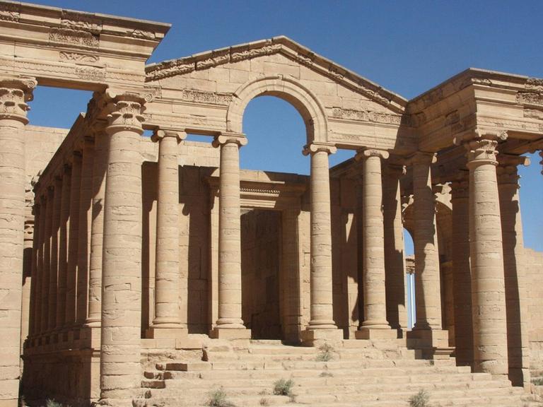 Ein antiker Tempel mit zwei Seitenflügeln und einer Treppe ohne Dach steht in der Wüste.