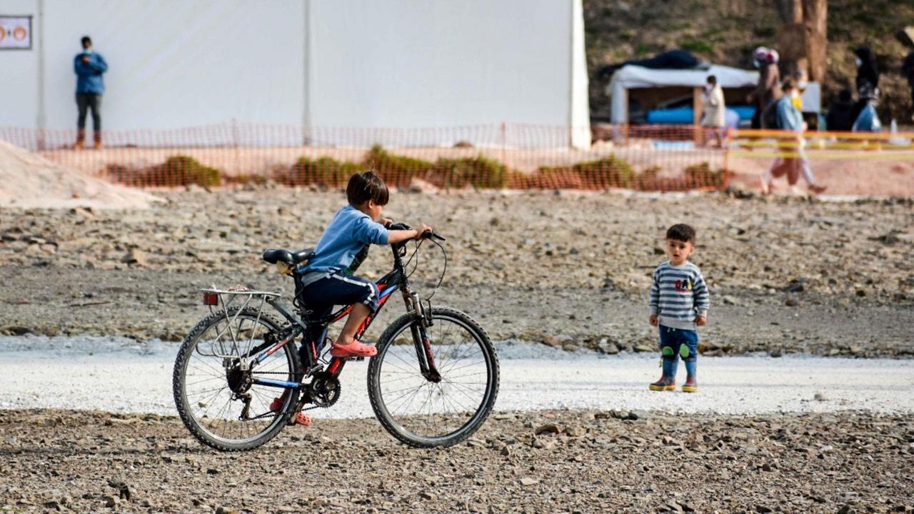 Ein Kind fährt vor Zelten auf einem für ihn zu großen Fahrrad, unweit steht ein kleineres Kind und schaut dabei zu Ein Kind fährt vor Zelten auf einem für ihn zu großen Fahrrad, unweit steht ein kleineres Kind und schaut dabei zu