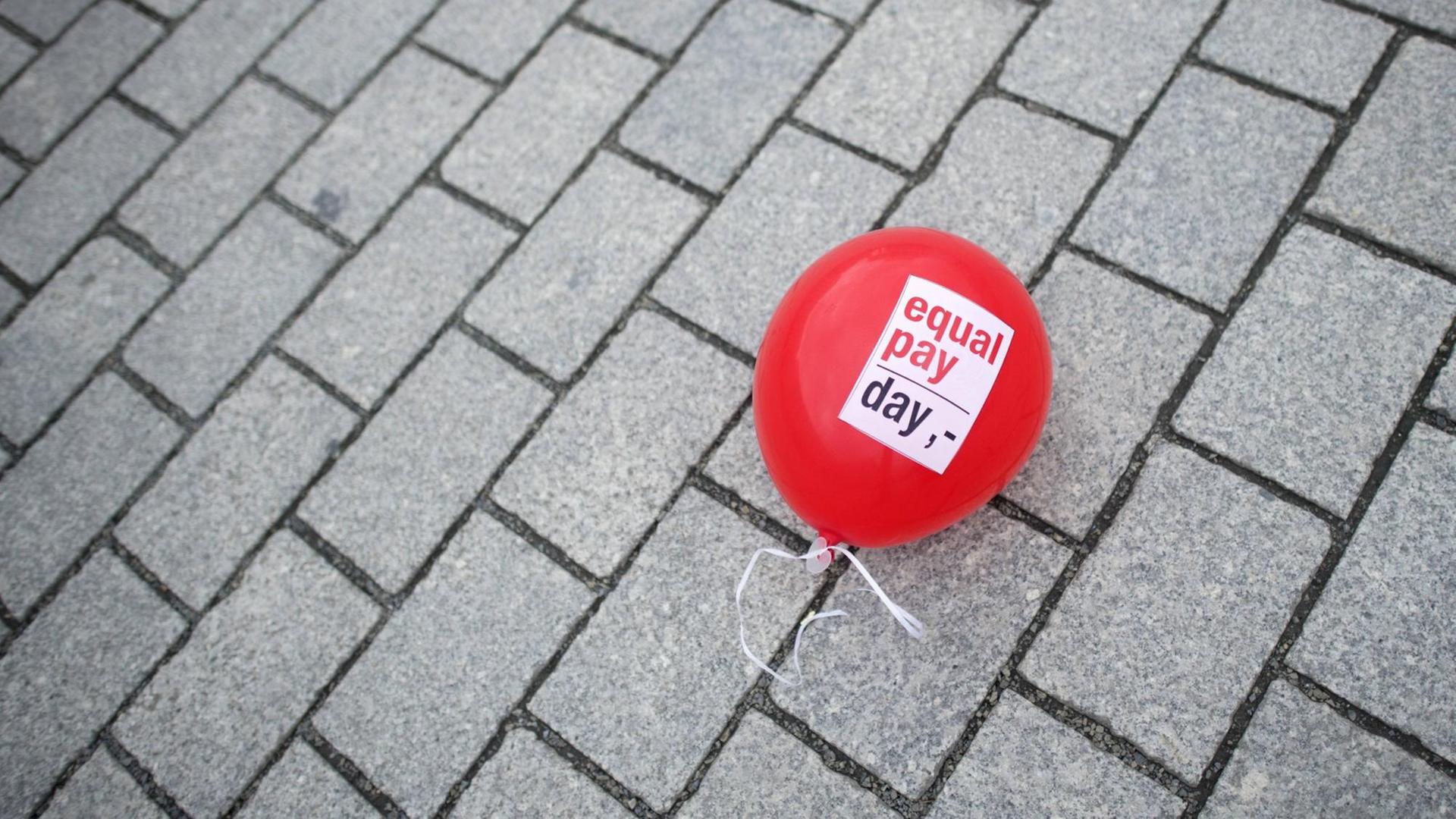 Ein roter Luftballon mit der Aufschrift "Equal Pay Day" liegt auf einem Pflasterboden. Ein roter Luftballon mit der Aufschrift "Equal Pay Day" liegt auf einem Pflasterboden.