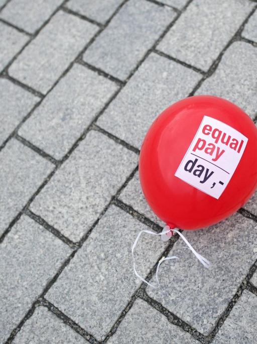 Ein roter Luftballon mit der Aufschrift "Equal Pay Day" liegt auf einem Pflasterboden.