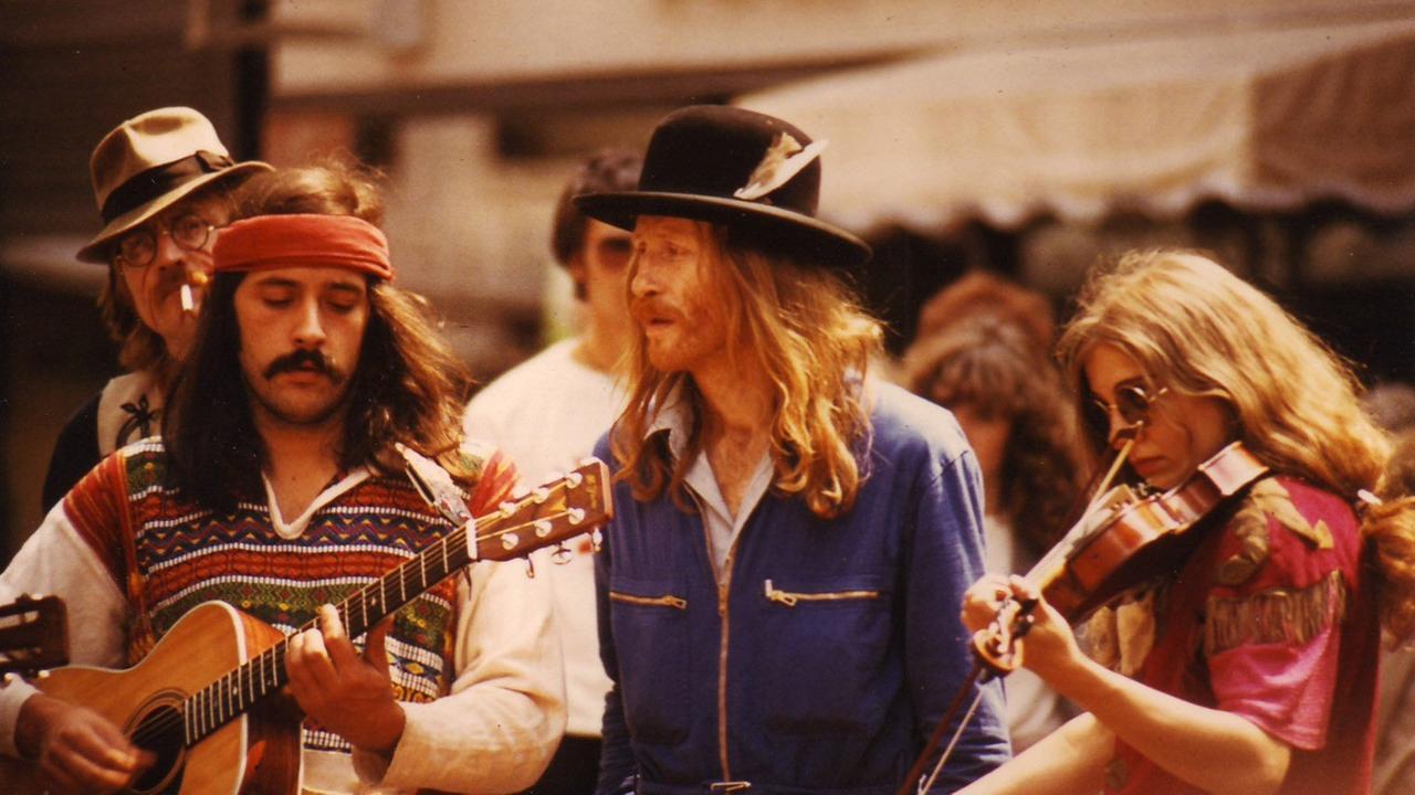Ein Mann spielt Gitarre und eine Frau spielt Geige. Zwischen den beiden steht ein Mann, der nach rechts guckt. Alle tragen Siebzigerjahre Outfits. Ein Mann spielt Gitarre und eine Frau spielt Geige. Zwischen den beiden steht ein Mann, der nach rechts guckt. Alle tragen Siebzigerjahre Outfits.