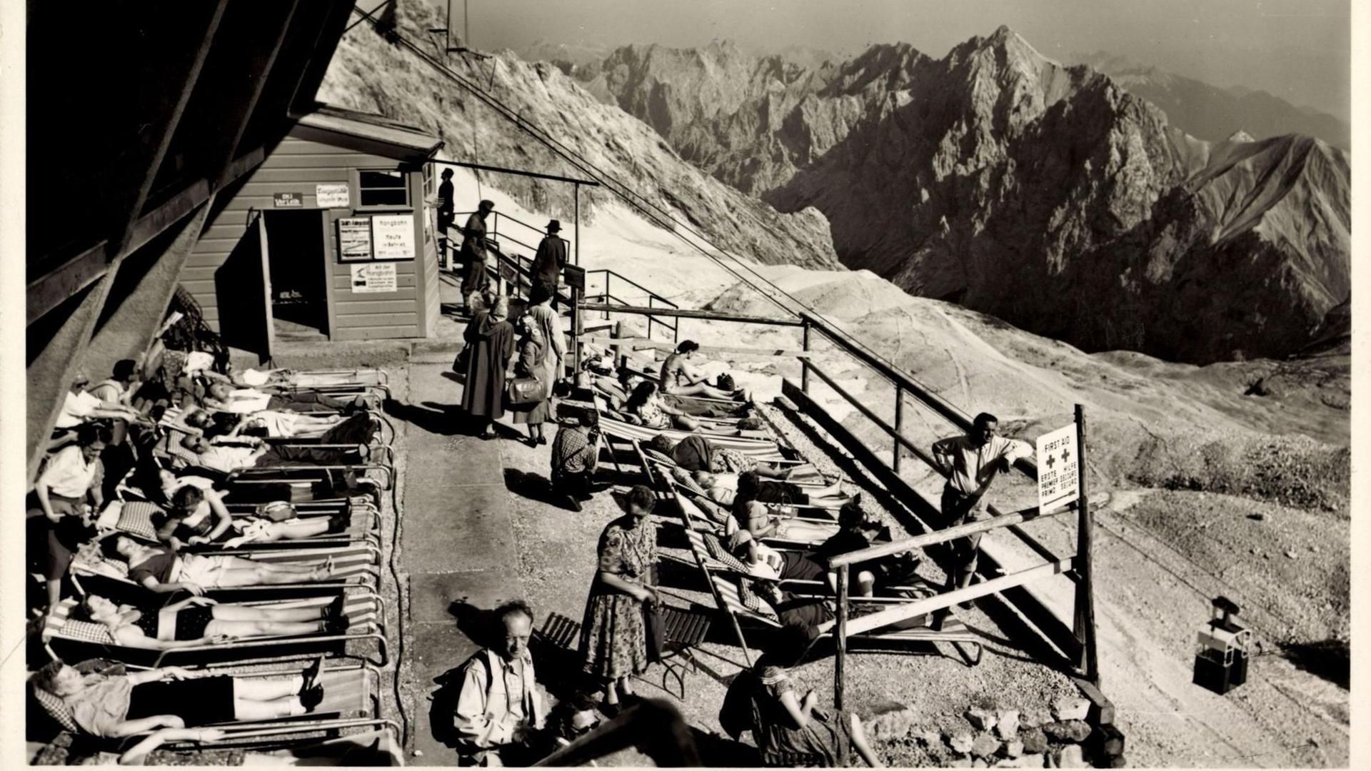 Ein Schwarzweiß-Foto zeigt etwa ein Dutzend Menschen in Liegestühlen sonnend auf einer mit Geländern eingefassten Terrasse. Im Hintergrund ein gewaltiges Alpen-Panorama