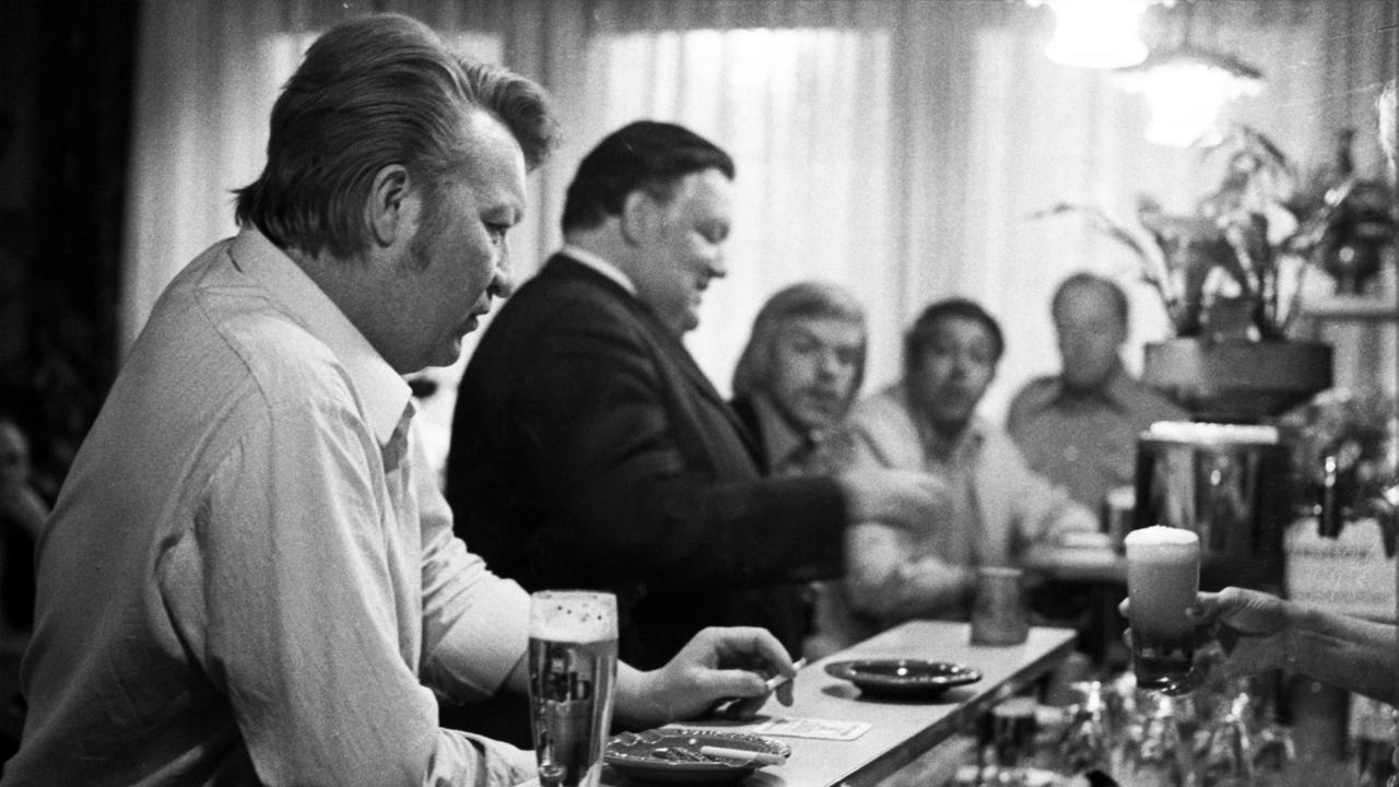 Fünf Männer sitzen in Dortmund an einer Theke und trinken Bier. Von rechts reicht eine Hand ein frisch gezapftes Bier.