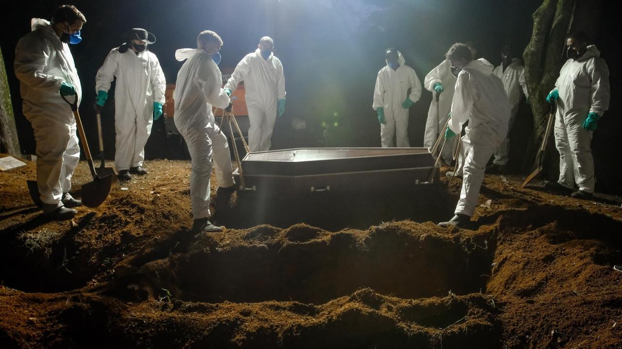 Mitarbeiter eines Friedhofs tragen einen Sarg, um eine an Covid-19 verstorbene Person zu beerdigen. Es ist Nacht, im Hintergrund sind Lichtstrahler aufgestellt, man sieht mehrere ausgehobene Gräber hintereinander. Mitarbeiter eines Friedhofs tragen einen Sarg, um eine an Covid-19 verstorbene Person zu beerdigen. Es ist Nacht, im Hintergrund sind Lichtstrahler aufgestellt, man sieht mehrere ausgehobene Gräber hintereinander.