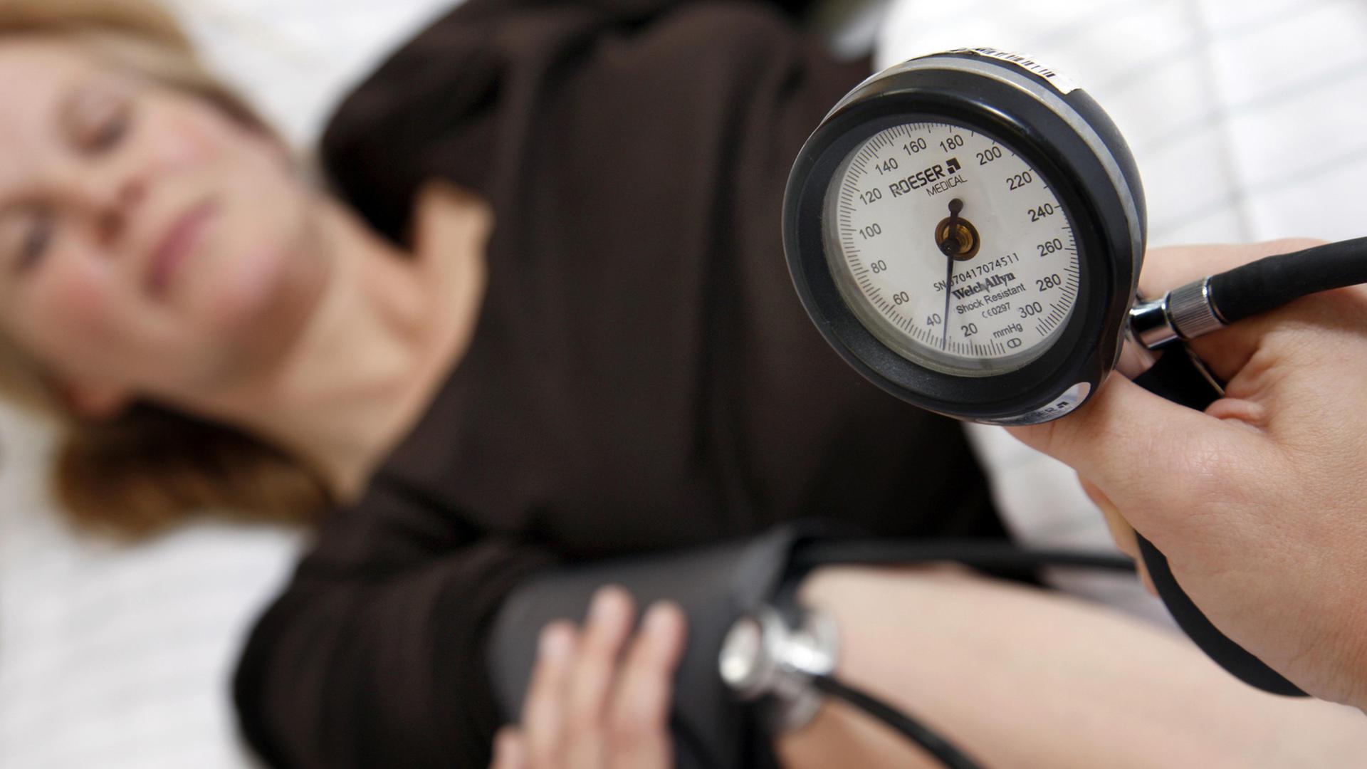 Ein Frau mit schwarzem Oberteil auf einer Liege; im Vordergrund ist die Messuhr eine Blutdruckgeräts zu sehen, das eine andere Person, die nicht im Bild ist, in der Hand hält.