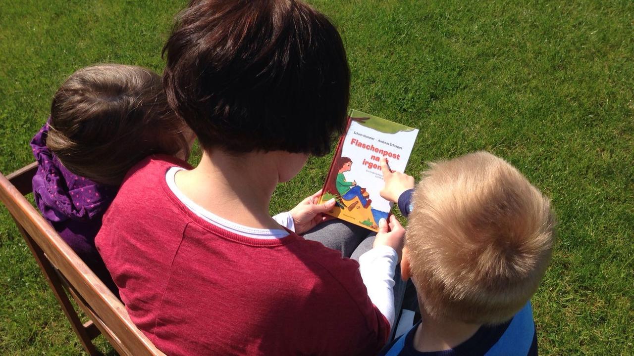 Eine Mutter sitzt mit ihren zwei Kindern auf einer Bank und hält ein Buch in den Händen. Eine Mutter sitzt mit ihren zwei Kindern auf einer Bank und hält ein Buch in den Händen.