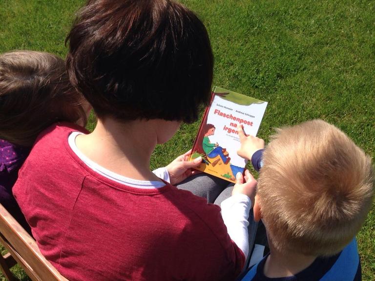 Eine Mutter sitzt mit ihren zwei Kindern auf einer Bank und hält ein Buch in den Händen.