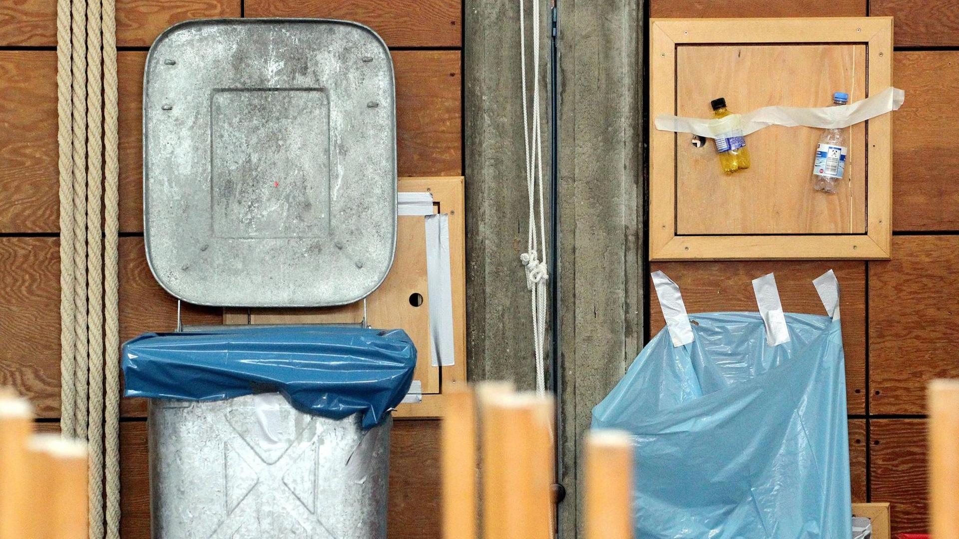 Eine Mülltonne steht neben einem an der Wand festgeklebten Müllsack, über dem zwei Plastikflaschen an der Wand hängen.