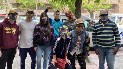 Eine Gruppe junger Leute mit Käppis und buntem Mundschutz posieren auf einer Straße für die Kamera.