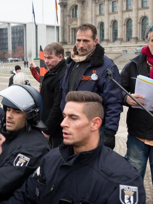 Vier Reichsbürger stehen hinter einem Mikrofoständer, einer von ihnen hält das Mikro. Drei Polizisten stehen davor, sie riegeln die Kundgebung gegen Gegendemonstranten ab.