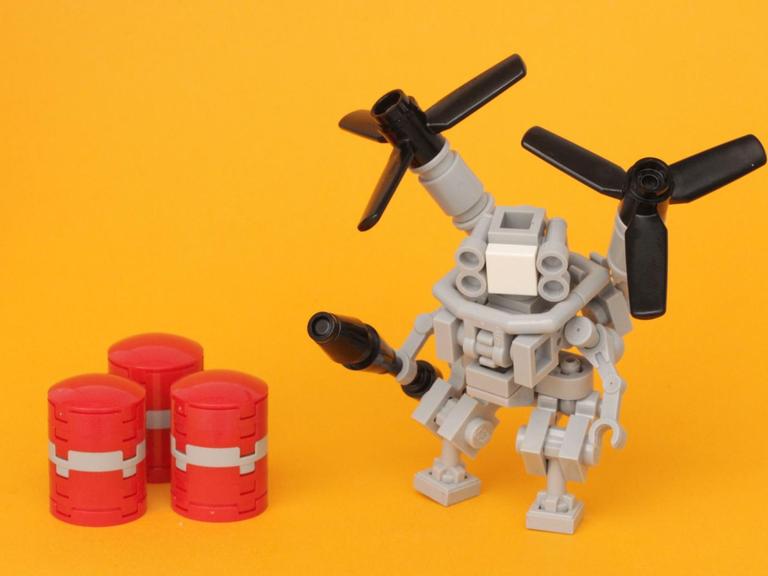 Lego-Roboter mit Propellern neben drei kleinen, roten Fässern vor einem gelben Hintergrund