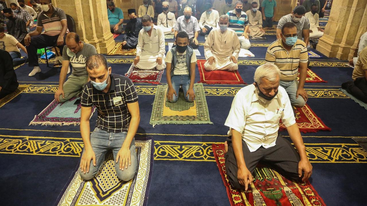 Männer knieen und beten in einer Moschee. Zwischen ihnen gibt es jeweils eine größere Lücke.