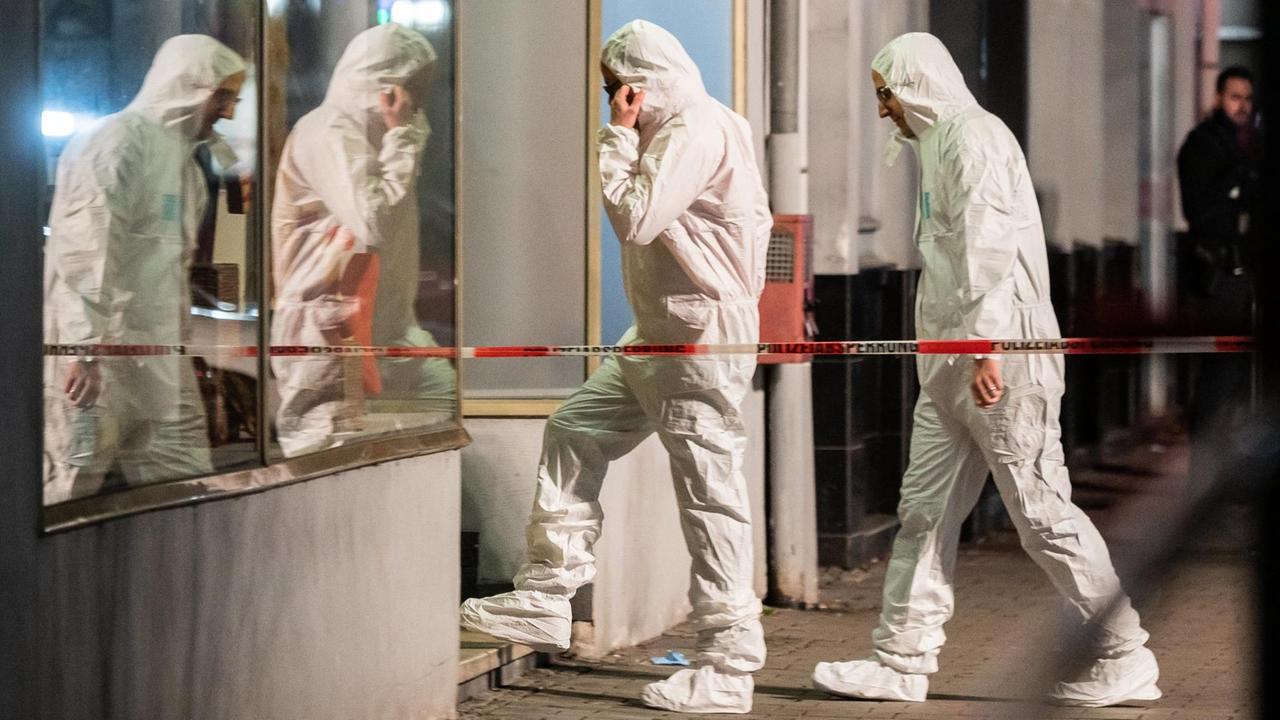 Zwei Männer in weißen Schutzanzügen betreten ein haus bei Dunkelheit. Sie spiegeln sich in einer Fensterscheibe. Im Vordergrund ein weiß-rotes Absperrband. Zwei Männer in weißen Schutzanzügen betreten ein haus bei Dunkelheit. Sie spiegeln sich in einer Fensterscheibe. Im Vordergrund ein weiß-rotes Absperrband.