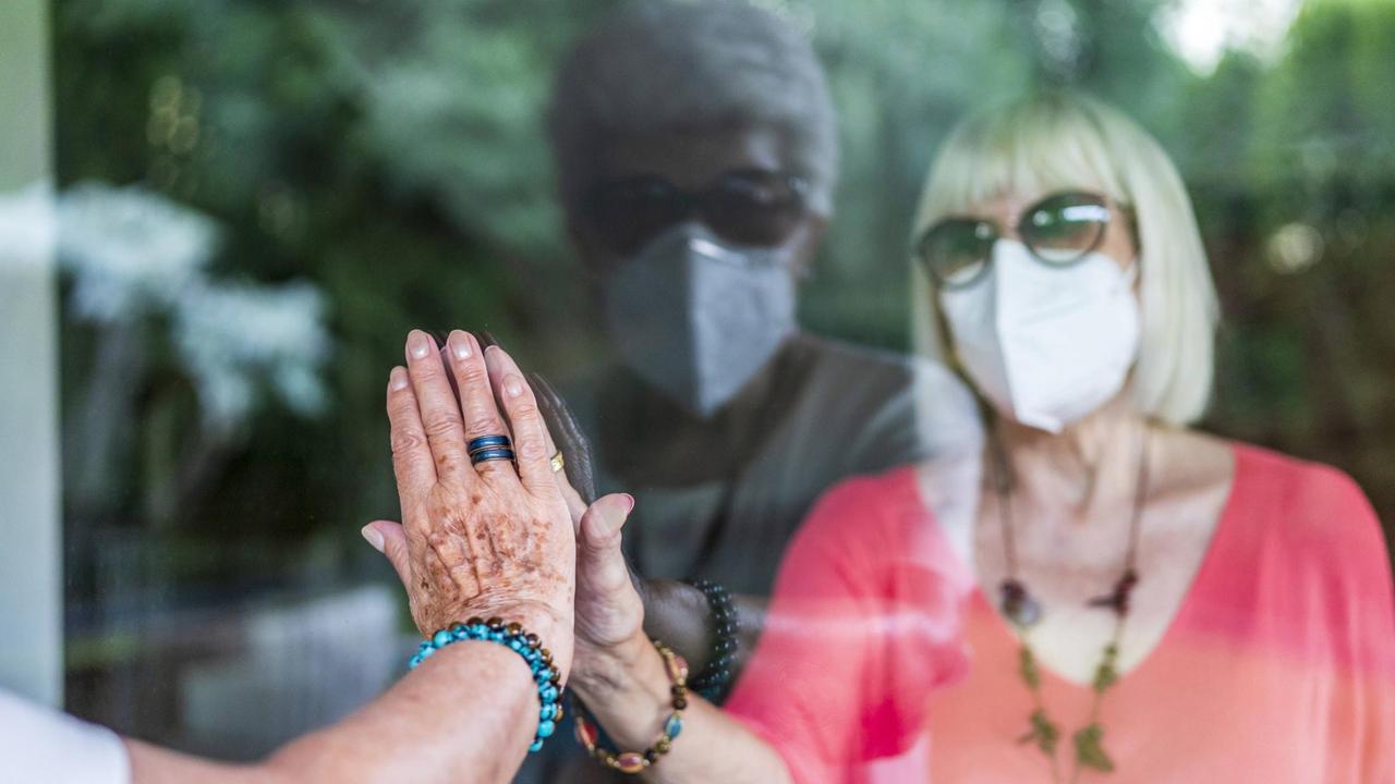 Die Hand einer Seniorin berührt durch eine Glasscheibe hindurch die Hand einer Frau, die hinter der Scheibe mit Maske steht.