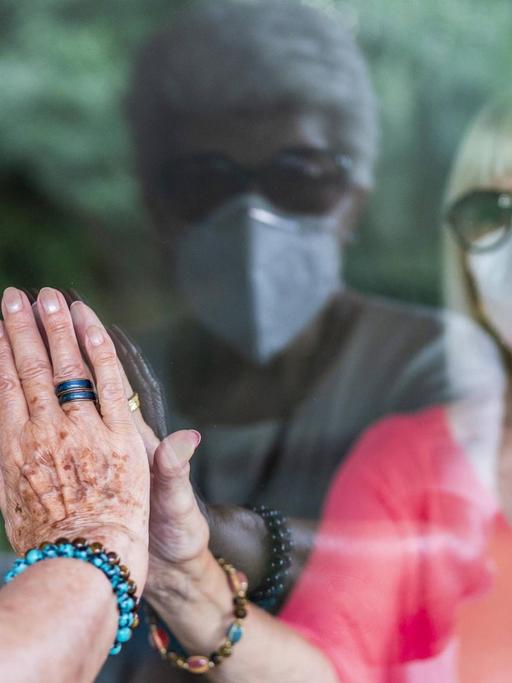 Die Hand einer Seniorin berührt durch eine Glasscheibe hindurch die Hand einer Frau, die hinter der Scheibe mit Maske steht.