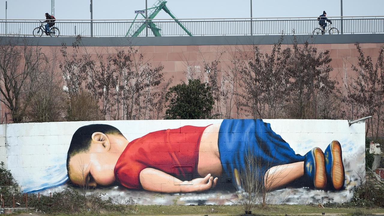 Das Foto des ertrunkenen syrischen Flüchtlingskindes Aylan Kurdi dient als Vorlage für ein gesellschaftskritisches Graffiti, das am 11.03.2016 auf der Osthafenmole in Frankfurt am Main (Hessen) zu sehen ist. Rund 40 Stunden haben die Künstler Justus Becker und Oguz Sen an dem rund 120 Quadratmeter großen Motiv gearbeitet. Foto: Arne Dedert/dpa | Verwendung weltweit Das Foto des ertrunkenen syrischen Flüchtlingskindes Aylan Kurdi dient als Vorlage für ein gesellschaftskritisches Graffiti, das am 11.03.2016 auf der Osthafenmole in Frankfurt am Main (Hessen) zu sehen ist. Rund 40 Stunden haben die Künstler Justus Becker und Oguz Sen an dem rund 120 Quadratmeter großen Motiv gearbeitet. Foto: Arne Dedert/dpa | Verwendung weltweit