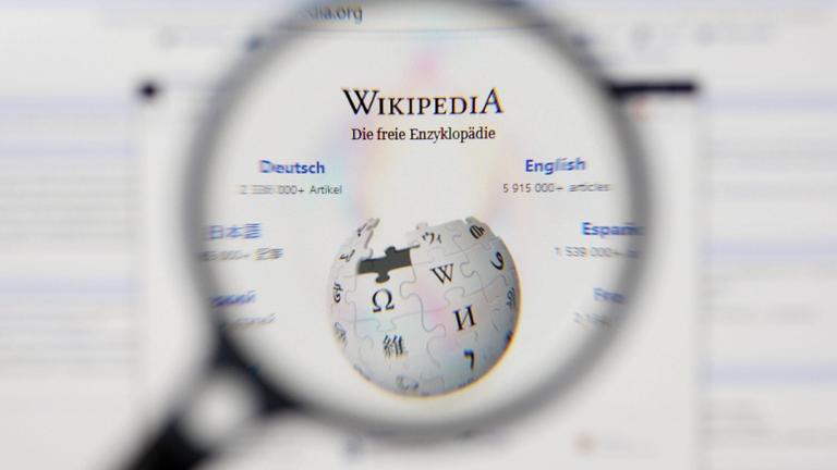 Eine Lupe vergrößert das Logo von 'Wikipedia - Die freie Enzyklopädie