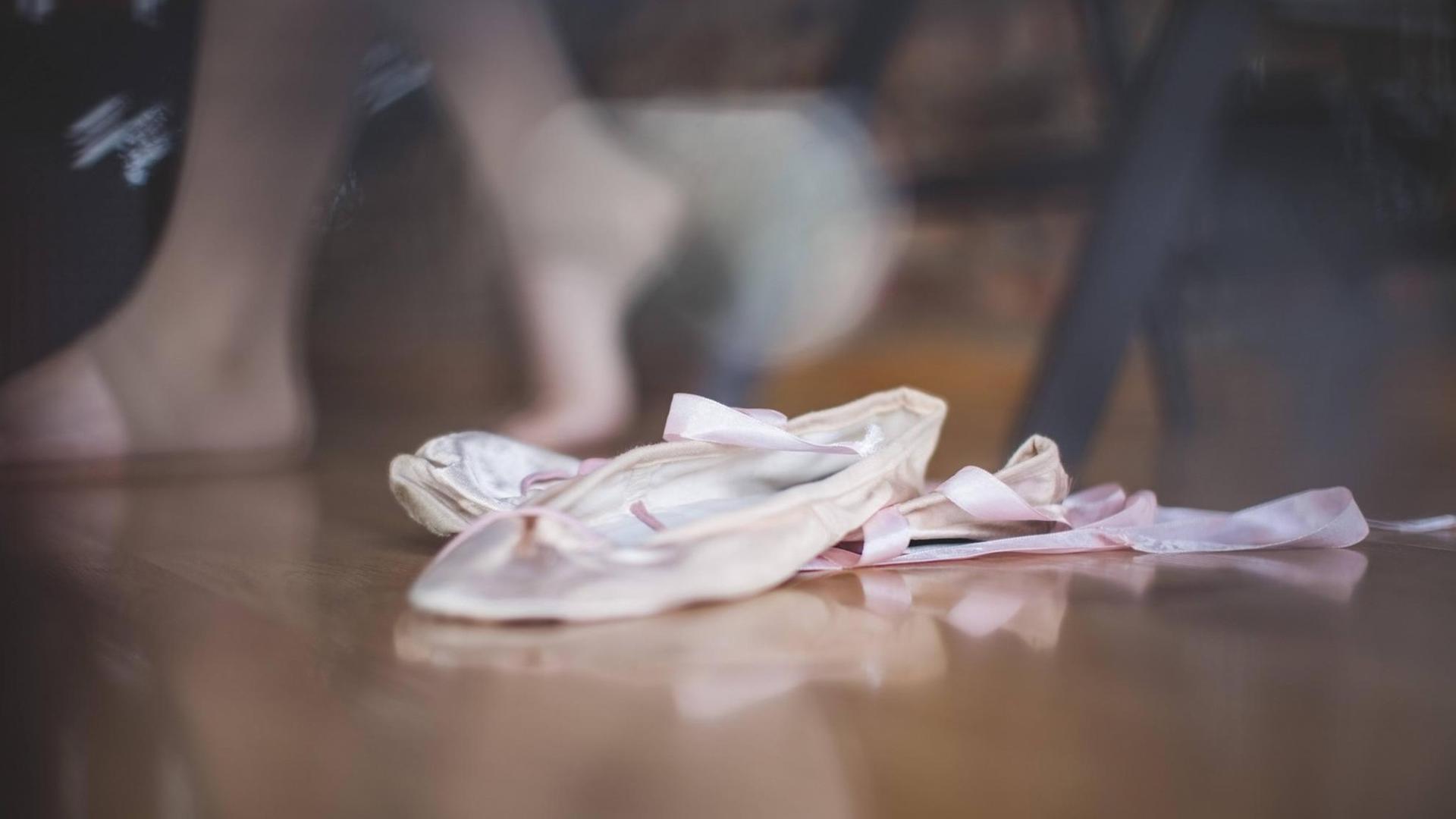 Das Foto zeigt ein paar helle Ballettschuhe, die etwas zerdrückt auf dem Boden eines Ballettsaals liegen. Das Foto zeigt ein paar helle Ballettschuhe, die etwas zerdrückt auf dem Boden eines Ballettsaals liegen.
