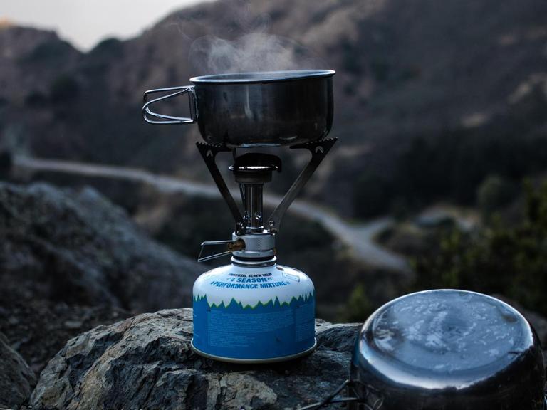 Ein kleiner Edelstahltopf steht auf einem Campingkocher, im Hintergrund sind Berge zu sehen.