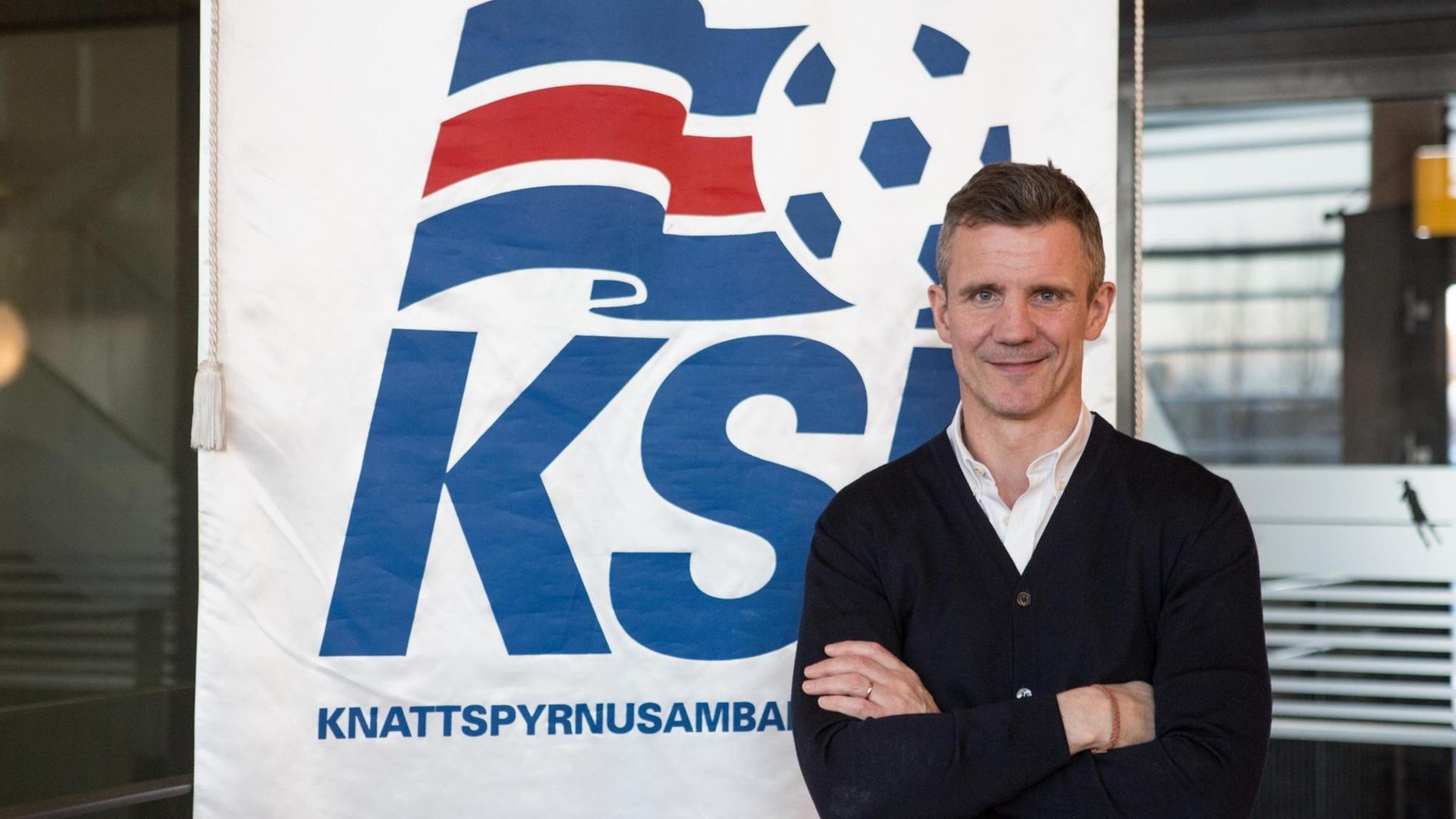 Guðni Bergsson steht mit verschränkten Armen vor einem Plakat des Verbandes KSÍ, er lächelt und trägt ein weißes Hemd und einen schwarzen Strickpulli Guðni Bergsson steht mit verschränkten Armen vor einem Plakat des Verbandes KSÍ, er lächelt und trägt ein weißes Hemd und einen schwarzen Strickpulli