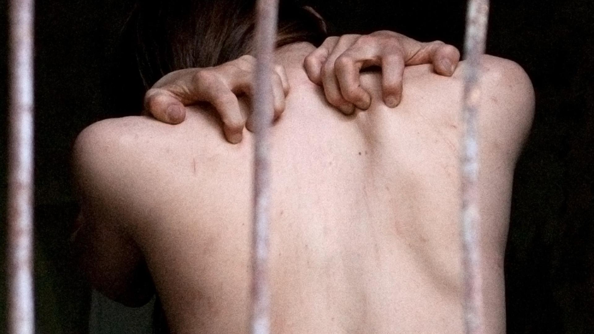 Hinter drei vertikalen Metallstangen ist ein nackter oberer Rückenausschnitt eines Menschen zu sehen, der seine Finger in die Schultern presst Hinter drei vertikalen Metallstangen ist ein nackter oberer Rückenausschnitt eines Menschen zu sehen, der seine Finger in die Schultern presst