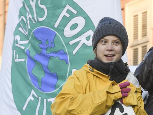 Die winterlich gekleidete Klimaaktivistin Greta Thunberg steht bei einer Kundgebung vor einer weißen Fahne, auf der das runde Logo "Fridays for Future" zu sehen ist. Im Hintergrund sind Häuser zu erkennen.