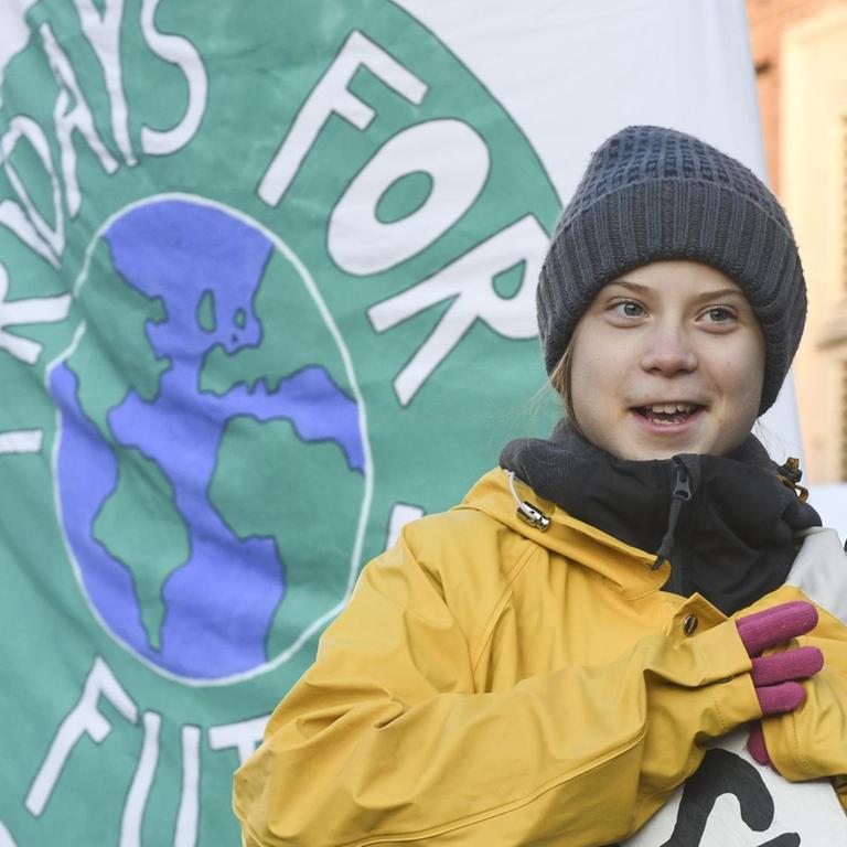Die winterlich gekleidete Klimaaktivistin Greta Thunberg steht bei einer Kundgebung vor einer weißen Fahne, auf der das runde Logo "Fridays for Future" zu sehen ist. Im Hintergrund sind Häuser zu erkennen.