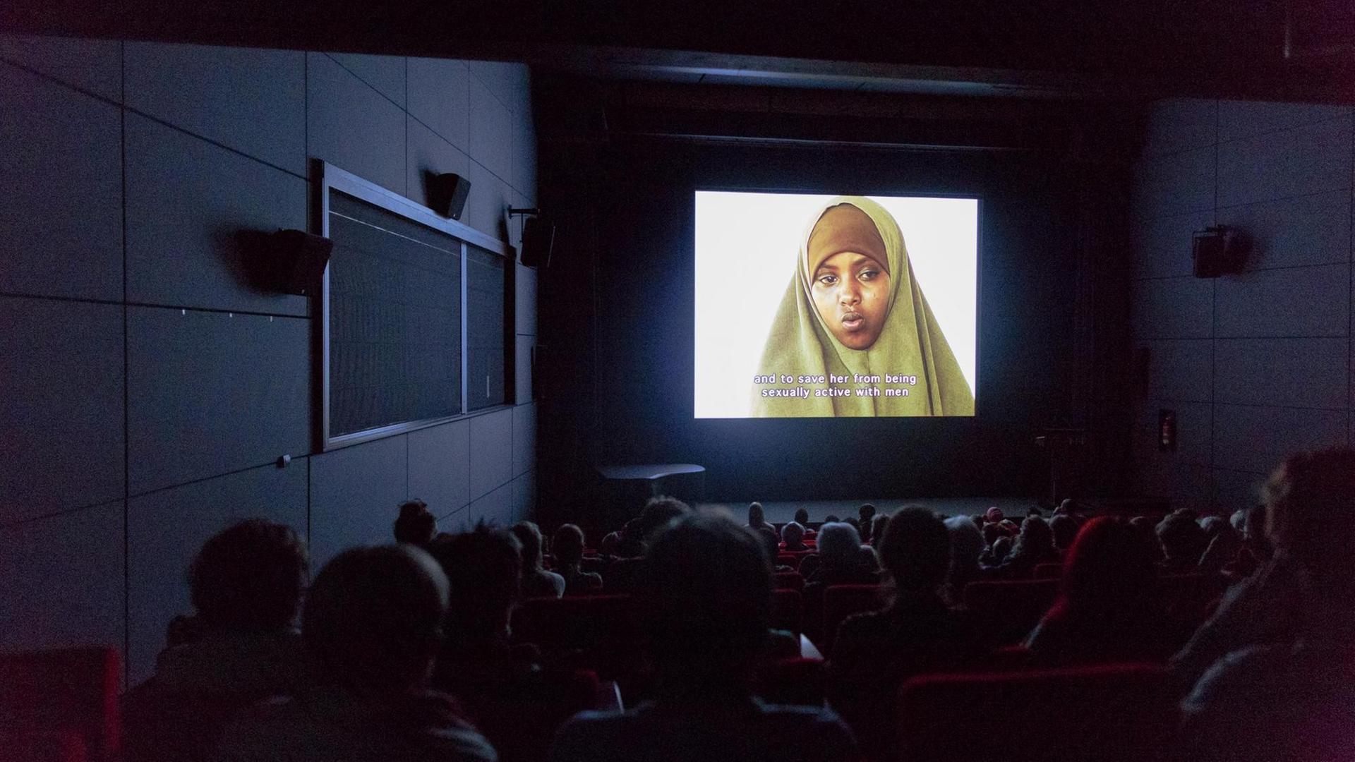 In einem dunklen Kinosaal schaut das Publikum auf die Leinwand - dort ist eine junge Frau mit Kopftuch zu sehen