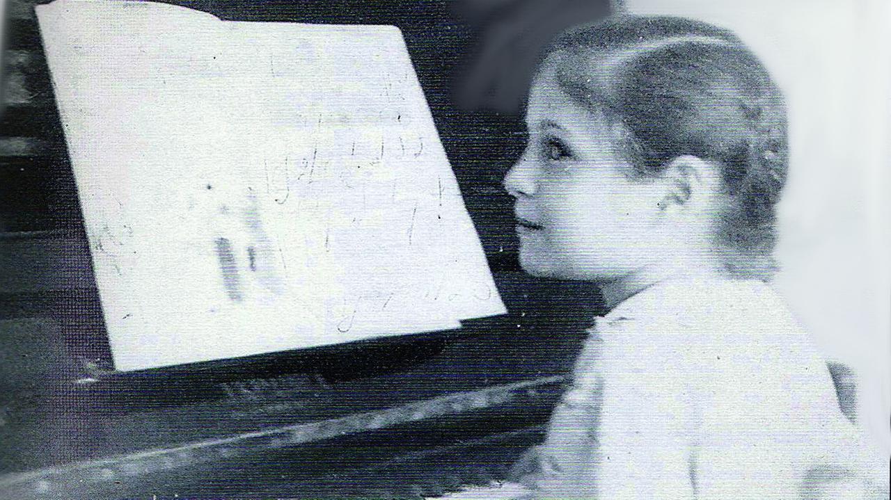 Ein junges Mädchen mit geflochtenen Zöpfen sitzt an einem Klavier, auf dem ein aufgeschlagenes Notenheft zu erkennen ist.
